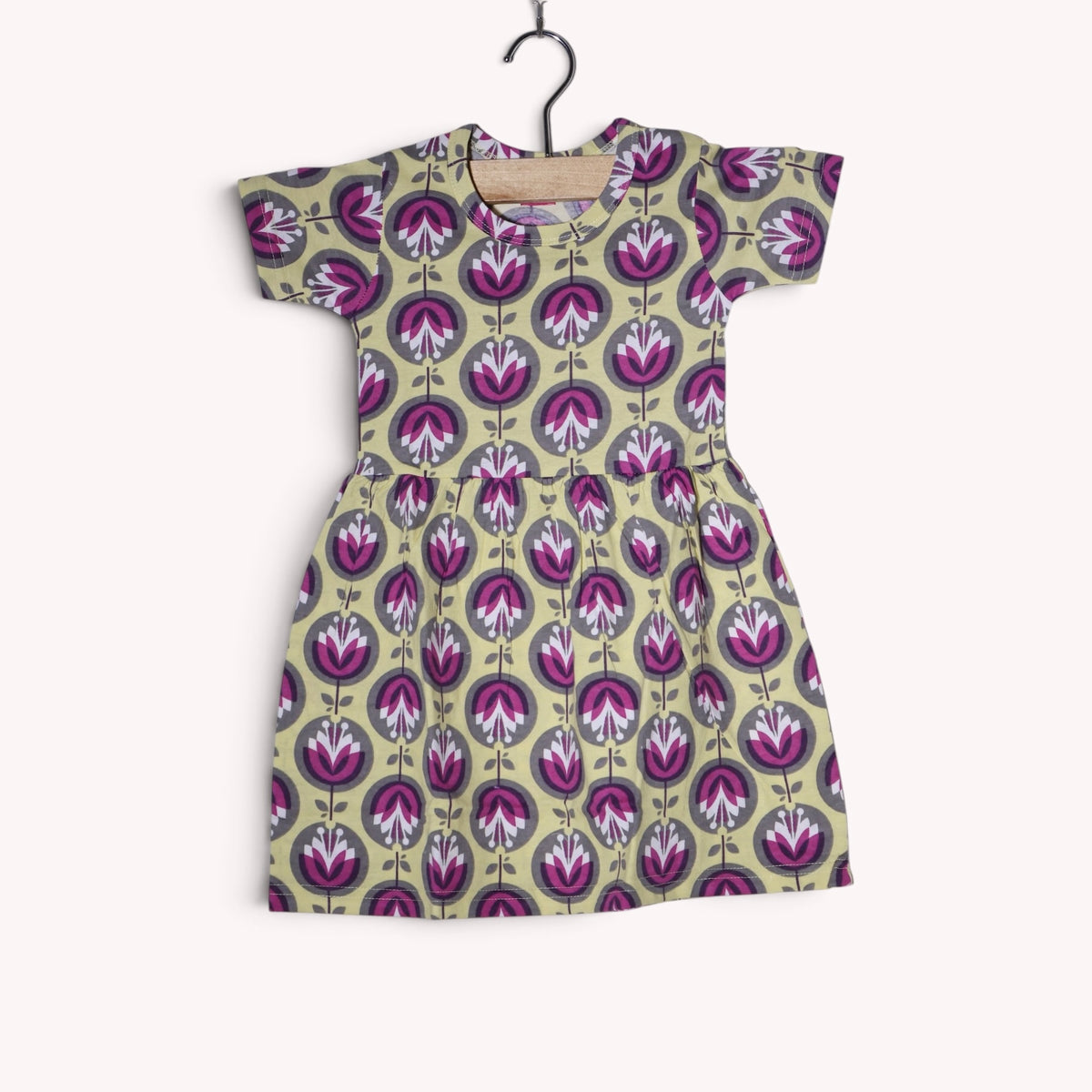 Dusty beige baby frock with a repeating stylised floral print in purple, magenta, white and grey, featuring short sleeves and a gathered waist