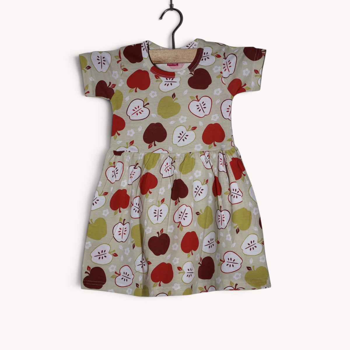 Dusty cream baby frock with an allover apple print in red, maroon, olive green and white, featuring short sleeves and a gathered waist