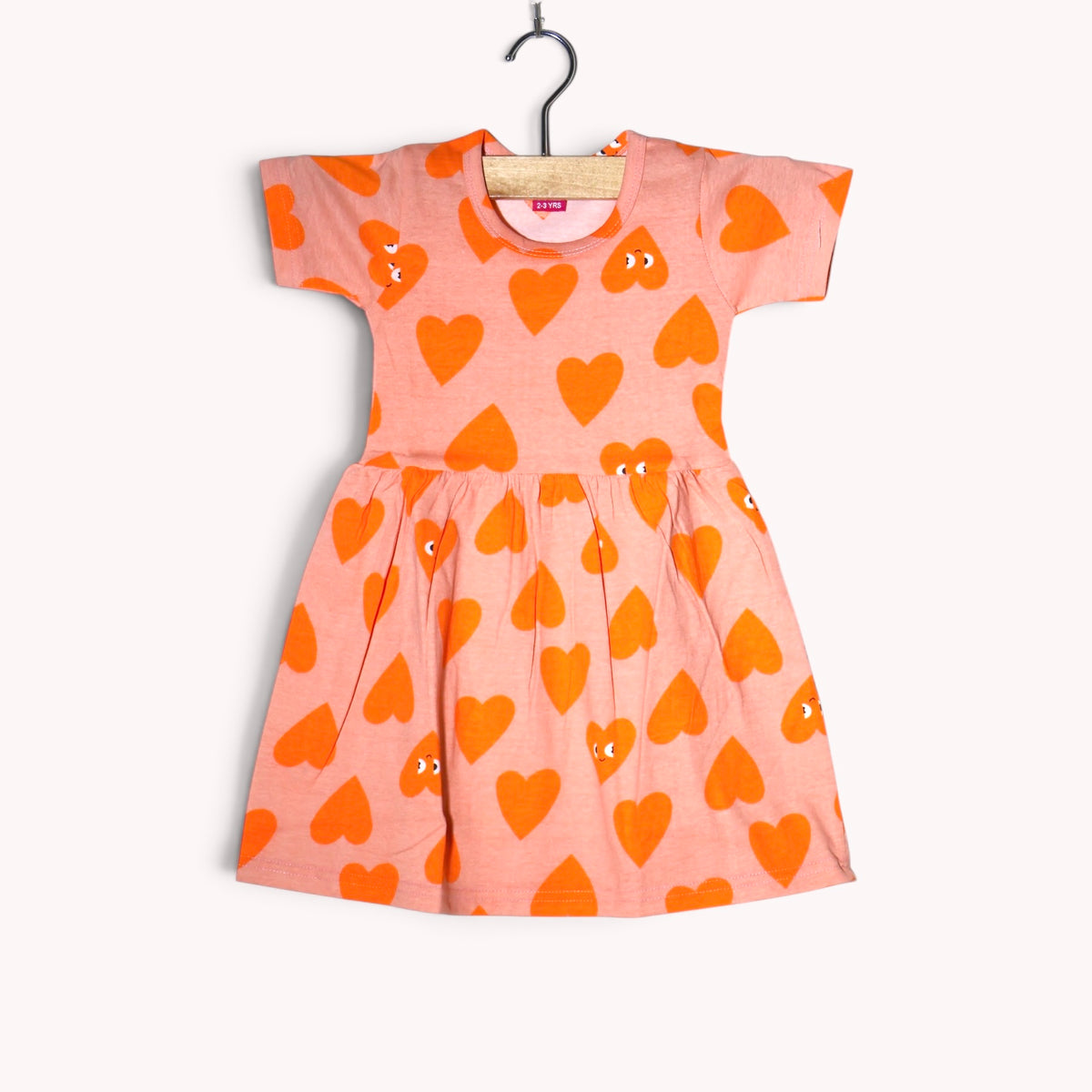Light peach cotton baby frock featuring a bright orange heart print with eye details and short sleeves.